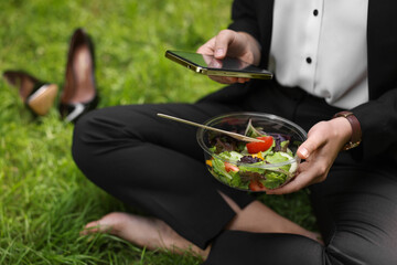 Lunch time. Businesswoman with container of salad using smartphone on green grass outdoors, closeup
