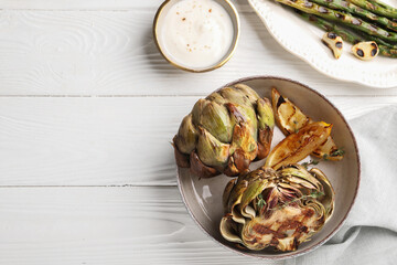Tasty grilled artichokes served with sauce and asparagus on white wooden table, flat lay. Space for text