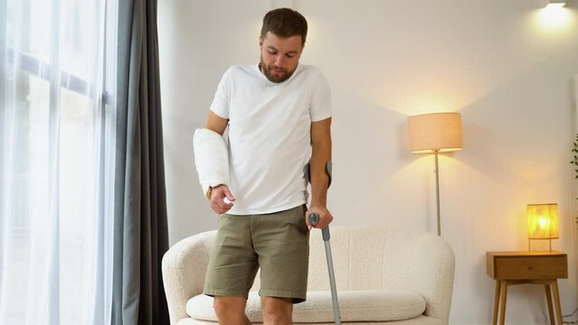 Happy man with broken arm and leg gets up with a crutch from sofa. Rehabilitation concept