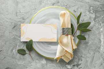 Blank invitation card, plate, napkin and eucalyptus leaves on grey table, flat lay. Space for text