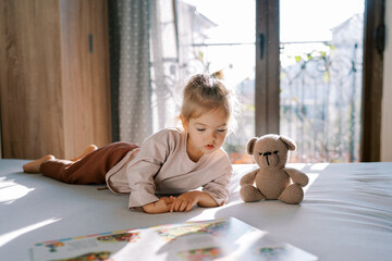 Little girl lies on her side on a bed with a teddy bear and looks at a picture book