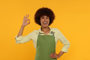 Happy young woman in apron showing ok gesture on orange background