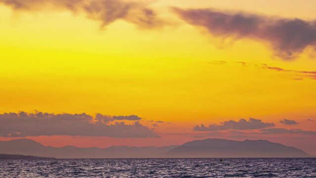 In Malaga, Spain, the captivating time lapse depicts a stunning marine landscape with majestic mountains, clouds, and the reflection of radiant shades of yellow sunlight.