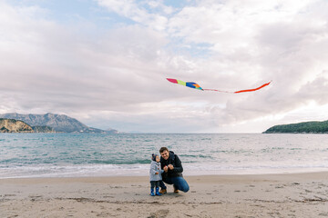 Dad, squatting down, launches a colorful kite by the sea with a little girl