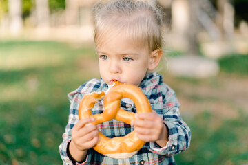 Little girl thoughtfully nibbles on a big pretzel holding it with both hands in the park
