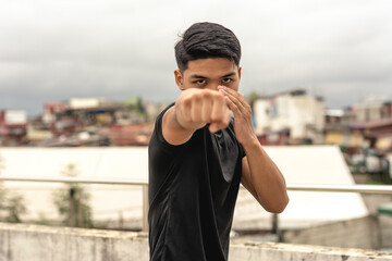 A young asian man makes a straight right hand while practicing his boxing skills on the rooftop of a building. Looking serious while doing shadowboxing training, gritty urban scene. - obrazy, fototapety, plakaty