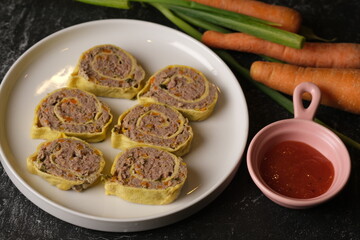 Rolade Daging Sapi. Beef roulade on a round white ceramic plate. Made from ground beef, pepper, nutmeg, eggs, garlic, carrot, onion leaves. Served with tomato sauce.