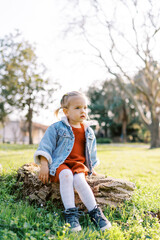Naklejka na ściany i meble Little girl sits on a stump in a sunny meadow and looks thoughtfully into the distance