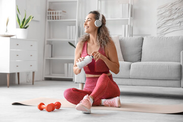 Smiling adult woman in sportswear with bottle of water sitting on yoga mat at home - Powered by Adobe