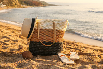 Stylish beach bag, coconut, hat and flip-flops on sand near sea