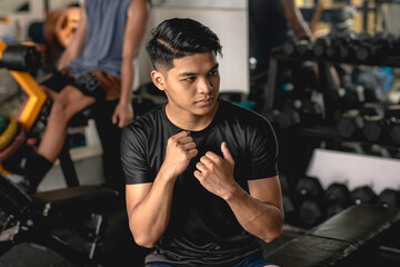 A young handsome asian man practicing his boxing combo skills while sitting on a bench at the gym. Muscle memory and reflex example. - obrazy, fototapety, plakaty