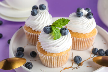 Plate of tasty cupcakes with blueberries on lilac background