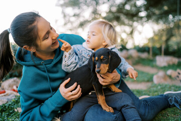 Smiling mother sitting with a puppy and a little daughter on her knees on the lawn - obrazy, fototapety, plakaty