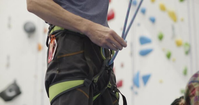 A sinewy armed climber preps to scale a rock climbing wall by first securing his harness with the proper knots.