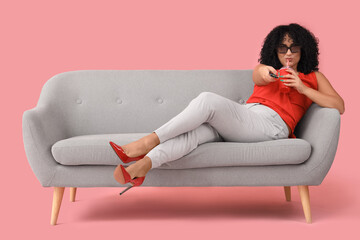 Beautiful woman in 3D glasses with soda watching TV on sofa against pink background