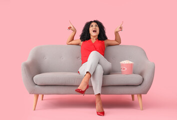 Beautiful woman with popcorn on sofa pointing at something against pink background - obrazy, fototapety, plakaty