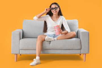 Young woman in 3D glasses with popcorn watching movie on sofa against yellow background