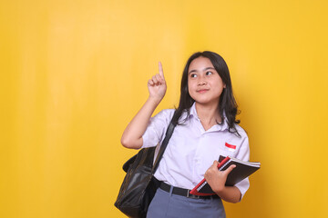 Cheerful Asian girl student pointing above to the empty side for educational ads or text while holding book, got an idea