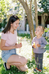 Smiling mom watches a little girl blowing soap bubbles through a stick with many holes