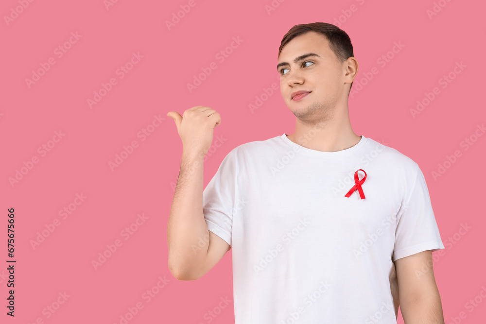 Sticker Handsome young man with red ribbon pointing at something on pink background. World AIDS day concept