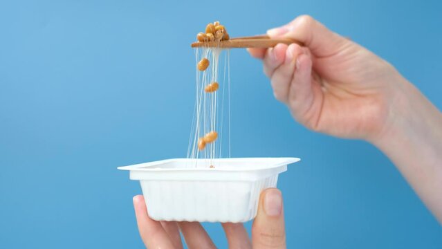 Female hand holding Japanese natto beans with wooden chopsticks on a blue background, side view.
