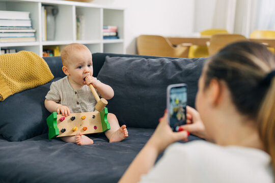 Mother is taking photo of her baby chewing on a wooden toy.