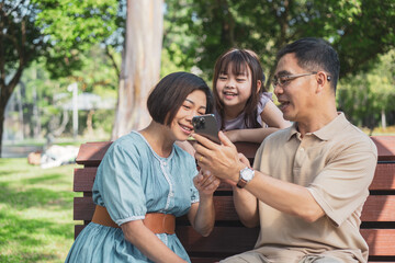 Cheerful Asian family of three having fun watching a phone together in the summer outdoors at the park.