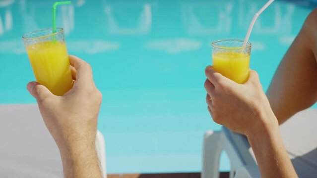Rear view hands of couple clinking glasses of orange juice when resting on poolside lounge chairs at resort hotel. Man and woman having drinks by the pool. Vacation, relaxation, recreation concept