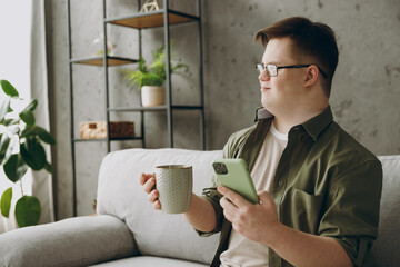 Young man with down syndrome wear glasses casual clothes use mobile cell phone drink tea sit on grey sofa couch stay at home flat rest spend free time in living room Genetic disease world day concept