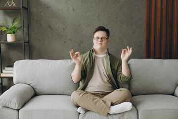 Young man with down syndrome wear glasses casual clothes meditate do yoga om gesture sits on grey sofa couch stay at home flat rest spend free time in living room. Genetic disease world day concept.