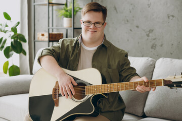 Young happy man with down syndrome wear glasses casual clothes play guitar look camera sits on grey...