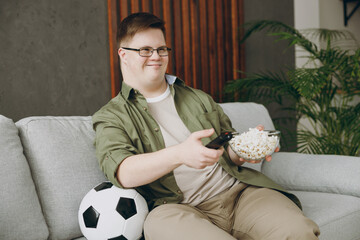 Young man with down syndrome wears glasses casual clothes watch tv football live stream sit on grey sofa couch stay at home flat rest spend free time in living room. Genetic disease world day concept