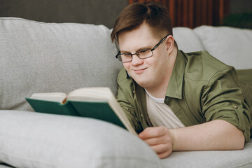 Side view young smart man with down syndrome wears glasses casual clothes reading book lay on grey sofa couch stay at home flat rest spend free time in living room. Genetic disease world day concept. - obrazy, fototapety, plakaty