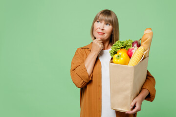 Elderly minded woman wear brown shirt casual clothes hold shopping paper bag with food products put hand prop up on chin, lost in thought isolated on plain green background Delivery service from shop