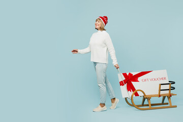 Full body merry elderly woman 50s years old wear sweater red hat posing carry gift certificate coupon voucher card for store on sled isolated on plain blue background. Happy New Year holiday concept. - obrazy, fototapety, plakaty