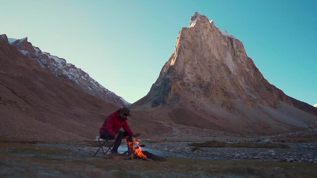 Indian male tourist sitting besides the bonfire in front of the Holy Gonbo Rangjon mountain in Zanskar Valley at Ladakh, India. Tourist warming himself with fire in cold Himalayas in India.