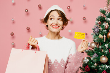 Merry young woman wear white sweater hat posing hold credit bank card package bags after shopping...