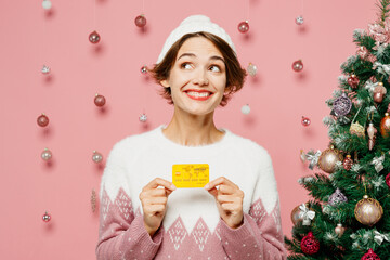 Merry wistful young woman wearing white sweater hat posing hold in hand mock up of credit bank card...