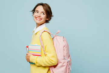 Side view young happy smiling woman student wearing casual clothes yellow sweater backpack bag hold books look aside on area isolated on plain blue background. High school university college concept.