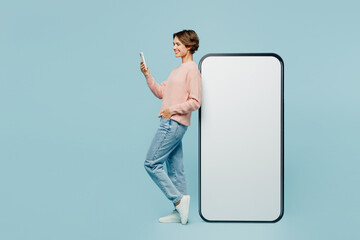 Full body side view young fun woman she wear beige knitted sweater casual clothes big huge blank screen mobile cell phone with workspace area use smartphone isolated on plain pastel blue background.