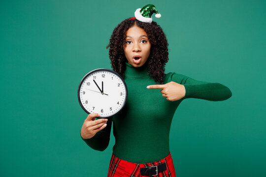 Merry shocked little kid teen girl wear hat casual clothes posing hold in hand point index finger on clock isolated on plain green background studio portrait. Happy New Year Christmas holiday concept.