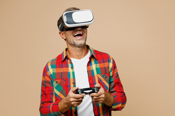 Fototapeta premium Adult man wear red shirt white t-shirt casual clothes watching in vr headset pc gadget hold play pc game with joystick console isolated on plain pastel light beige color background. Lifestyle concept.