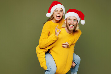 Fototapeta premium Merry young couple two friends man woman wear sweater Santa hat posing giving piggyback ride to joyful, sit on back show ok isolated on plain green background Happy New Year Christmas holiday concept