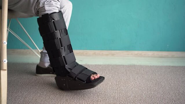 An injured man with an ankle brace orthosis for walking walks at home with a crutch