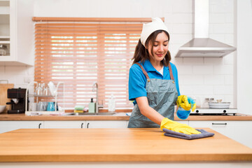 Professional Asian woman cleaning service wearing yellow rubber gloves, using a rag to wipe with spraying liquid detergent on the wooden top kitchen counter at home. Housekeeping cleanup, cleaner. - obrazy, fototapety, plakaty