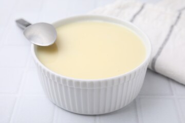 Bowl with condensed milk and spoon on white tiled table, closeup