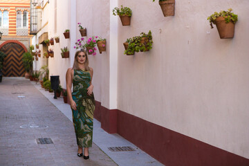 Pretty young blonde woman in green dress walks along a street decorated with flower pots and...
