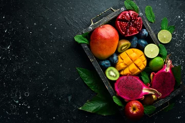 Tuinposter Tropical fruits in a wooden box: mango, dragon fruit, lime, pomegranate, plum, apple. On a black stone background. © Yaruniv-Studio