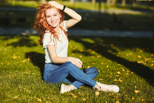 A lovely red-haired girl, warmed by the rays of the sun, is sitting on a lawn and posing for the camera. The girl is wearing a T-shirt with jeans, glasses on her face, and a modern gadget on her arm