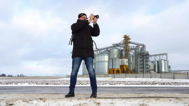 A photographer takes photos against the background of the agro-industrial complex. A man with a camera in winter in warm clothes on the background of a field covered with snow. silver silo 